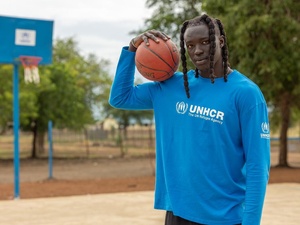 uN hombre con rastas en el cabello que viste una camiseta azul de ACNUR, sostiene una pelota de baloncesto color naranja en una cancha de baloncesto