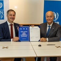 Dos hombres sonrientes que están sentados a una mesa sostienen un documento firmado en el que se lee “Acuerdo de colaboración conjunta entre el PNUD y ACNUR: 2026-2030”. Detrás de ellos se ven pancartas del PNUD y de ACNUR